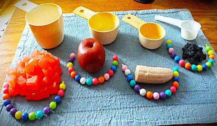 Colorful beads arranged around fruit and measuring cups on a blue surface.