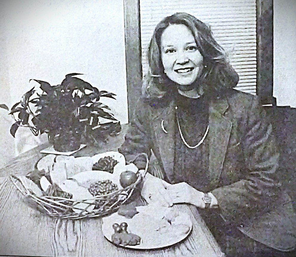 A woman smiling next to a plant and a basket of fruits.